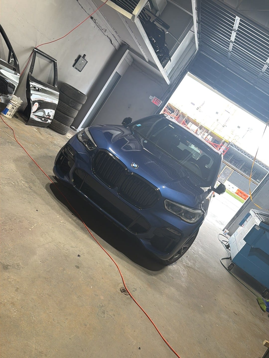 A blue BMW X5 inside the ABE Auto Collision bay, next to the open roll-up door